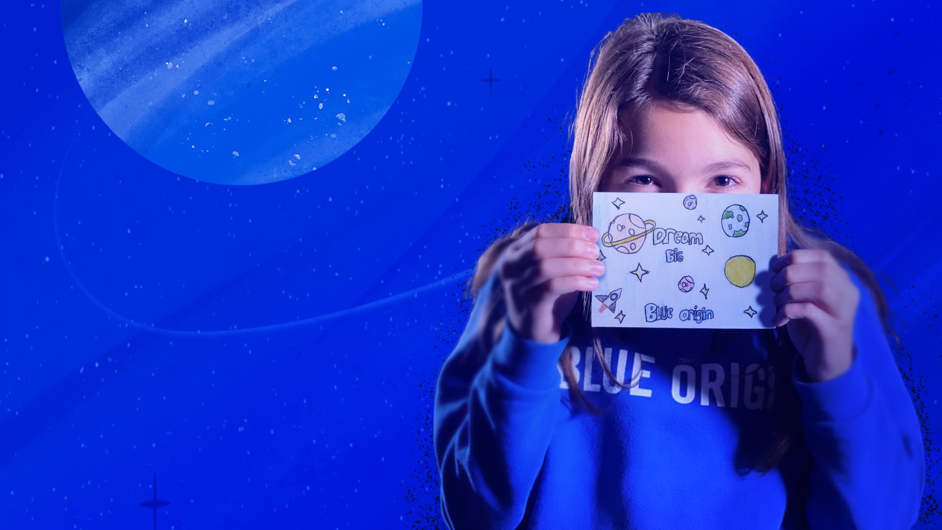 A girl in a 'Blue Origin' sweatshirt holds a paper postcard up to her face